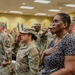 A formal military ceremony is taking place in an indoor venue, likely a conference or assembly room. Multiple U.S. Army soldiers in OCP (Operational Camouflage Pattern) uniforms are saluting while civilians stand with hands over their hearts, suggesting the playing of the National Anthem or Pledge of Allegiance. Various military unit flags are visible in the background.