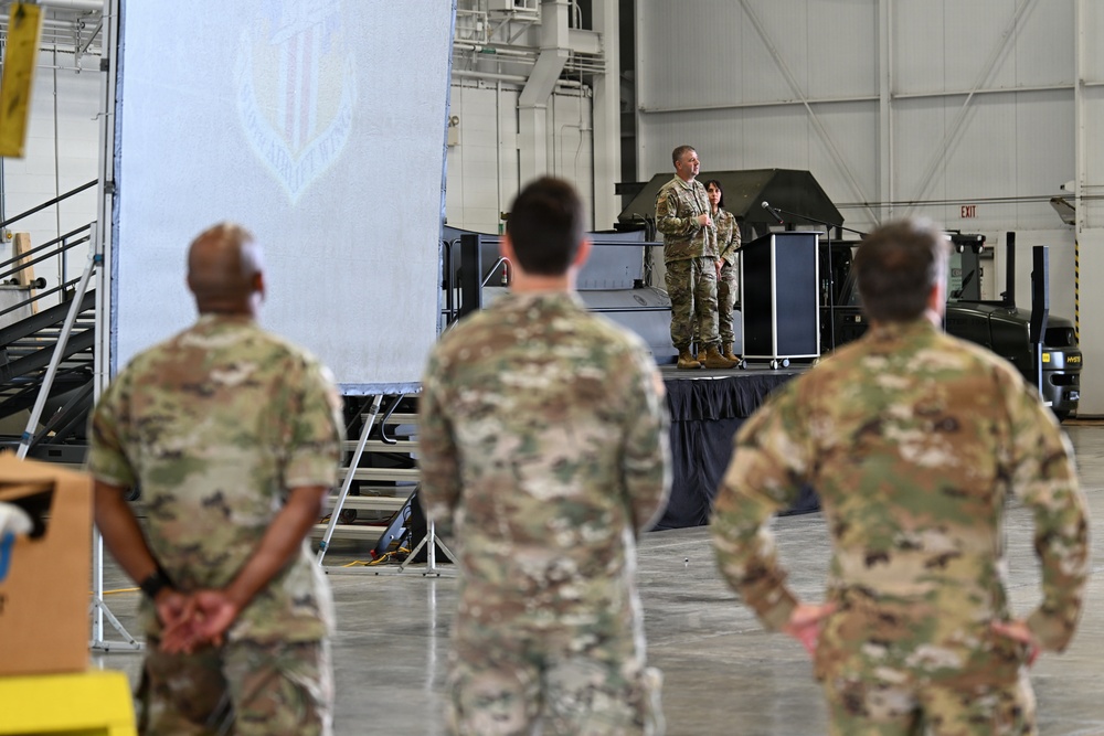A military briefing or ceremony is taking place inside a large hangar facility. Two uniformed personnel stand on a raised stage/platform addressing an audience of fellow service members in multicam/OCP camouflage uniforms. A large banner or screen with a military insignia is visible in the background.