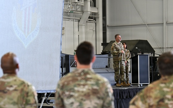 A military briefing or ceremony is taking place inside a large hangar facility. Two uniformed personnel stand on a raised stage/platform addressing an audience of fellow service members in multicam/OCP camouflage uniforms. A large banner or screen with a military insignia is visible in the background.