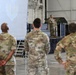 A military briefing or ceremony is taking place inside a large hangar facility. Two uniformed personnel stand on a raised stage/platform addressing an audience of fellow service members in multicam/OCP camouflage uniforms. A large banner or screen with a military insignia is visible in the background.