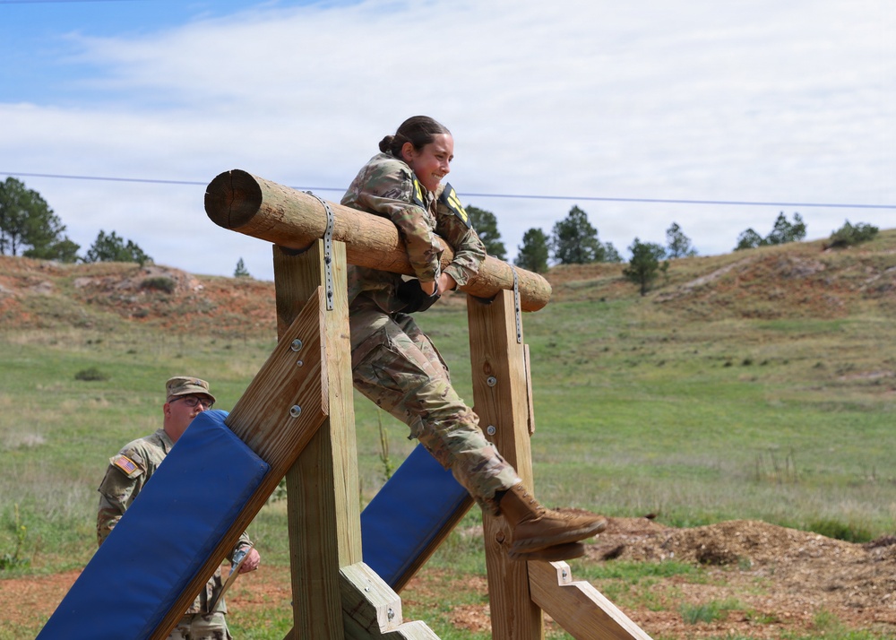 A soldier in US Army camouflage uniform is navigating a military obstacle course, specifically climbing over a horizontal log barrier supported by wooden posts. A second soldier in the background appears to be observing or waiting their turn. The setting is an outdoor training area with grassy hills and pine trees in the background.