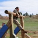 A soldier in US Army camouflage uniform is navigating a military obstacle course, specifically climbing over a horizontal log barrier supported by wooden posts. A second soldier in the background appears to be observing or waiting their turn. The setting is an outdoor training area with grassy hills and pine trees in the background.
