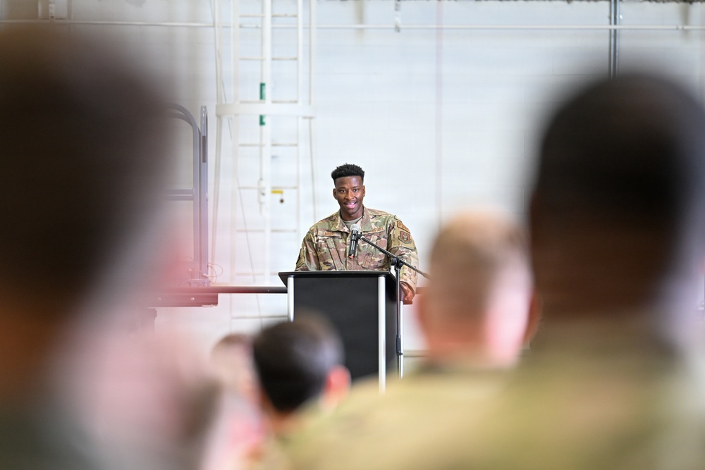 A military service member in camouflage uniform stands at a podium with a microphone, appearing to deliver a speech or address to an audience. The setting appears to be inside a large military hangar or facility. Several uniformed personnel are visible in the foreground as audience members, photographed from behind with shallow depth of field.
