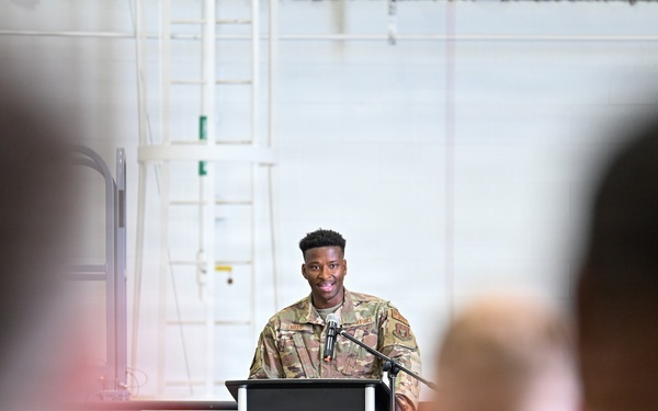 A military service member in camouflage uniform stands at a podium with a microphone, appearing to deliver a speech or address to an audience. The setting appears to be inside a large military hangar or facility. Several uniformed personnel are visible in the foreground as audience members, photographed from behind with shallow depth of field.