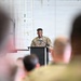 A military service member in camouflage uniform stands at a podium with a microphone, appearing to deliver a speech or address to an audience. The setting appears to be inside a large military hangar or facility. Several uniformed personnel are visible in the foreground as audience members, photographed from behind with shallow depth of field.