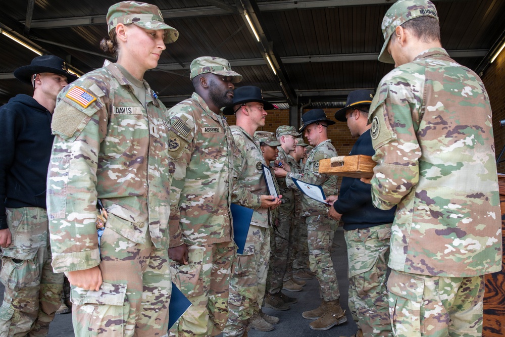 A group of U.S. Army soldiers in OCP (Operational Camouflage Pattern) uniforms are gathered in what appears to be an awards or recognition ceremony. Several soldiers are holding blue folders and documents, while one soldier holds a wooden presentation box, suggesting medals or coins are being awarded. The setting appears to be an indoor/covered facility such as a motor pool or hangar.
