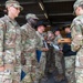 A group of U.S. Army soldiers in OCP (Operational Camouflage Pattern) uniforms are gathered in what appears to be an awards or recognition ceremony. Several soldiers are holding blue folders and documents, while one soldier holds a wooden presentation box, suggesting medals or coins are being awarded. The setting appears to be an indoor/covered facility such as a motor pool or hangar.