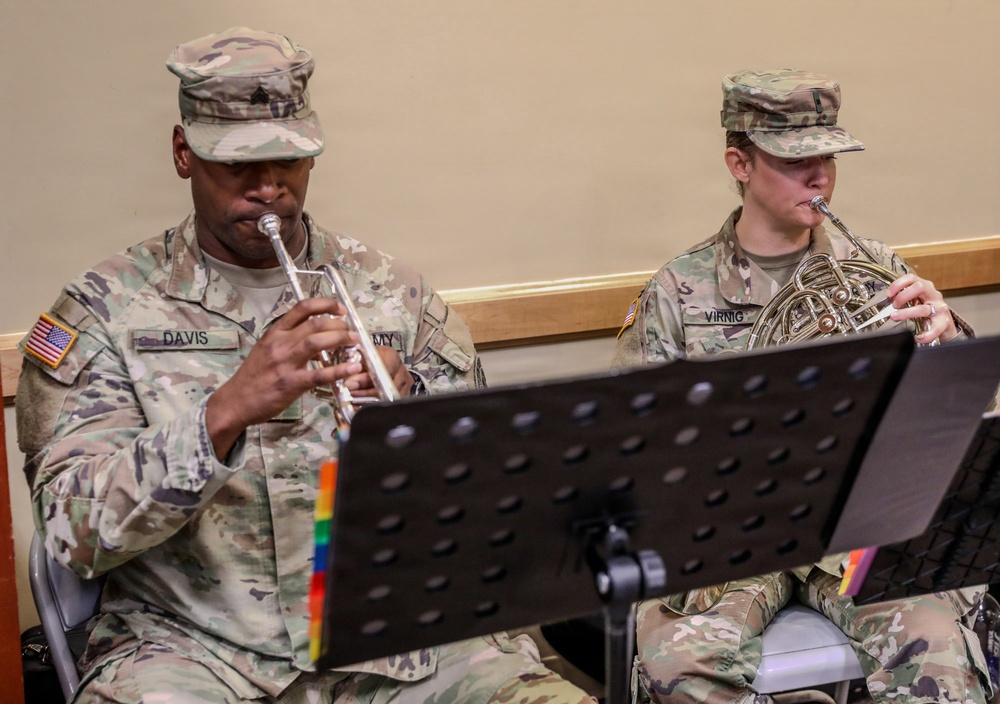Two U.S. Army soldiers in OCP (Operational Camouflage Pattern) uniforms are performing music from sheet music stands. The soldier on the left, identified by name tape as 'DAVIS,' is playing a trumpet, while the soldier on the right, identified as 'VIRNIG,' is playing a French horn. Both are seated and reading from music stands with colorful tab markers.