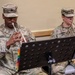 Two U.S. Army soldiers in OCP (Operational Camouflage Pattern) uniforms are performing music from sheet music stands. The soldier on the left, identified by name tape as 'DAVIS,' is playing a trumpet, while the soldier on the right, identified as 'VIRNIG,' is playing a French horn. Both are seated and reading from music stands with colorful tab markers.