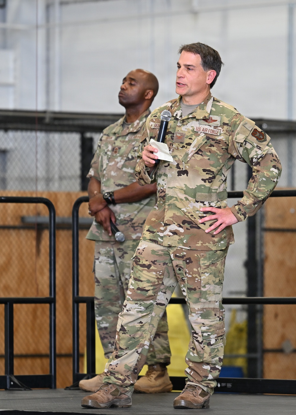 Two U.S. Air Force personnel in Operational Camouflage Pattern (OCP) uniforms are standing on a stage or platform, likely inside a hangar or large military facility. The individual in the foreground is speaking into a microphone while holding notes, suggesting an all-hands meeting, briefing, or official address. A second service member stands behind and to the left, also holding a microphone at his side.