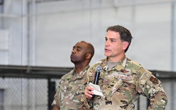 Two U.S. Air Force personnel in Operational Camouflage Pattern (OCP) uniforms are standing on a stage or platform, likely inside a hangar or large military facility. The individual in the foreground is speaking into a microphone while holding notes, suggesting an all-hands meeting, briefing, or official address. A second service member stands behind and to the left, also holding a microphone at his side.