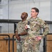 Two U.S. Air Force personnel in Operational Camouflage Pattern (OCP) uniforms are standing on a stage or platform, likely inside a hangar or large military facility. The individual in the foreground is speaking into a microphone while holding notes, suggesting an all-hands meeting, briefing, or official address. A second service member stands behind and to the left, also holding a microphone at his side.