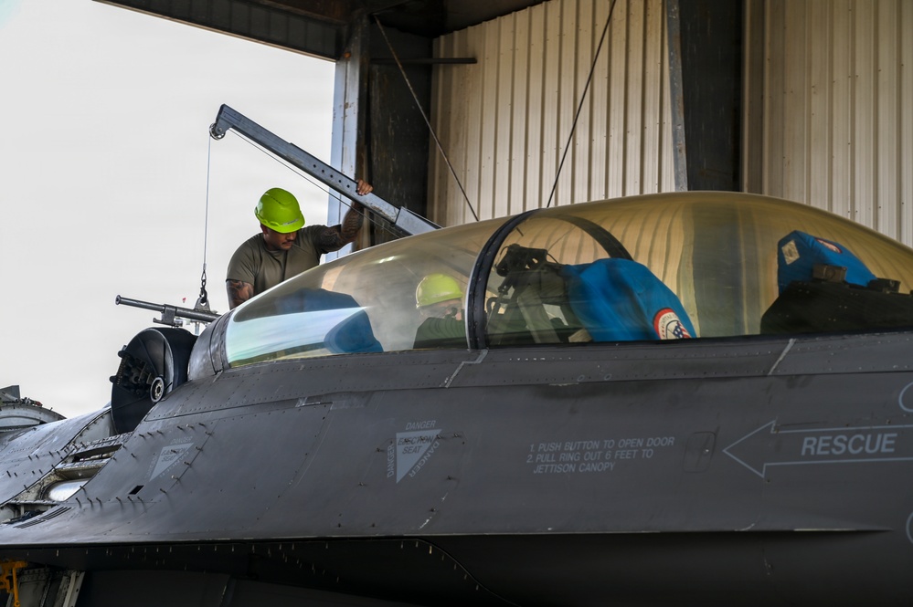A maintenance technician wearing a green hard hat and olive drab shirt is working on the canopy area of an F-16 Fighting Falcon jet fighter inside a hangar. The aircraft's cockpit canopy is open and covered with a gold-tinted protective cover, while blue protective covers are also visible on other components. The fuselage displays standard rescue markings and ejection seat warning labels.