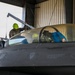 A maintenance technician wearing a green hard hat and olive drab shirt is working on the canopy area of an F-16 Fighting Falcon jet fighter inside a hangar. The aircraft's cockpit canopy is open and covered with a gold-tinted protective cover, while blue protective covers are also visible on other components. The fuselage displays standard rescue markings and ejection seat warning labels.