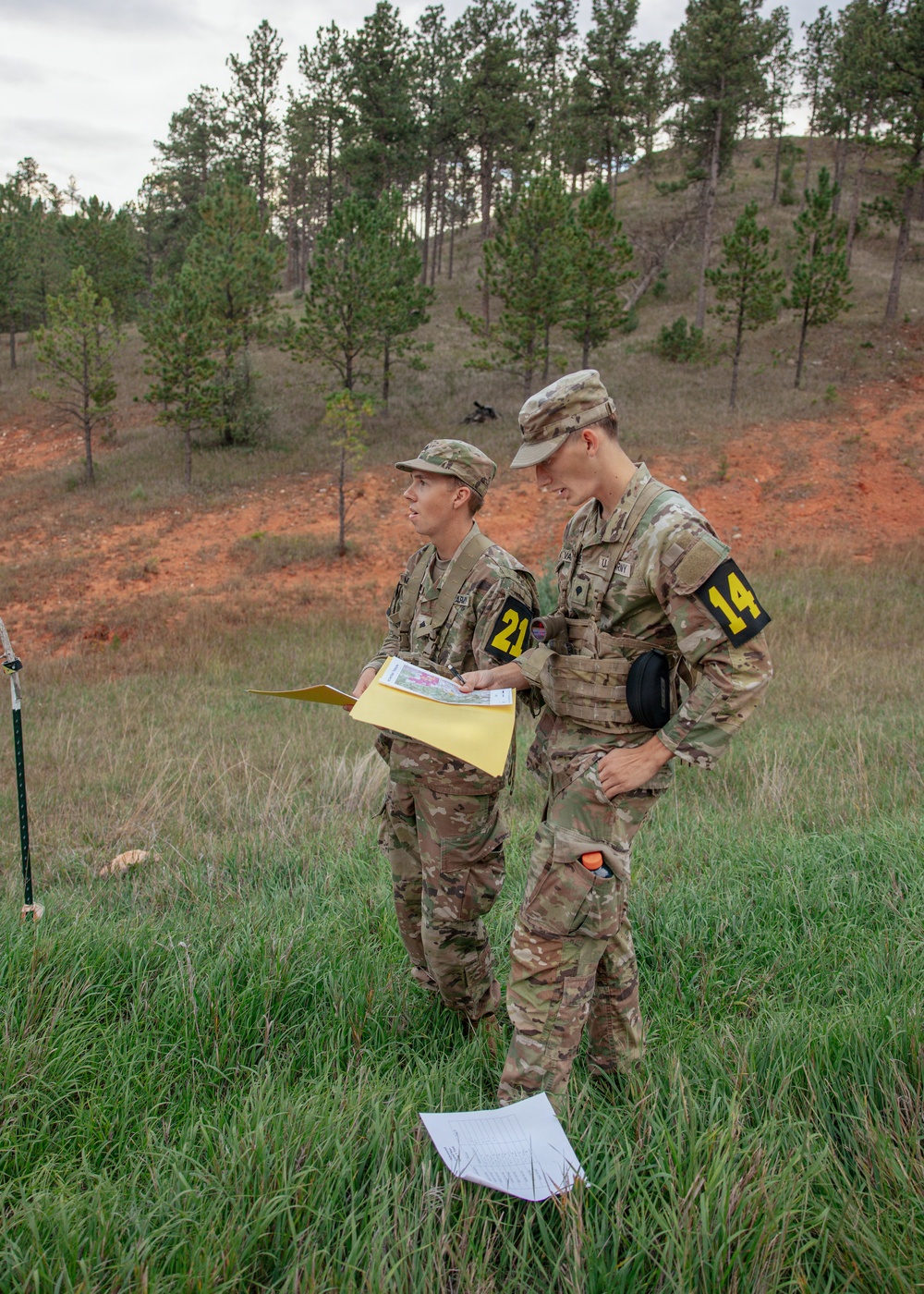 Two military personnel in OCP (Operational Camouflage Pattern) uniforms are standing in a grassy field consulting a map or document. They are wearing numbered armbands (21 and 14), suggesting participation in a land navigation exercise or military competition. The setting appears to be a training area with pine trees and red clay terrain in the background.