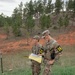 Two military personnel in OCP (Operational Camouflage Pattern) uniforms are standing in a grassy field consulting a map or document. They are wearing numbered armbands (21 and 14), suggesting participation in a land navigation exercise or military competition. The setting appears to be a training area with pine trees and red clay terrain in the background.