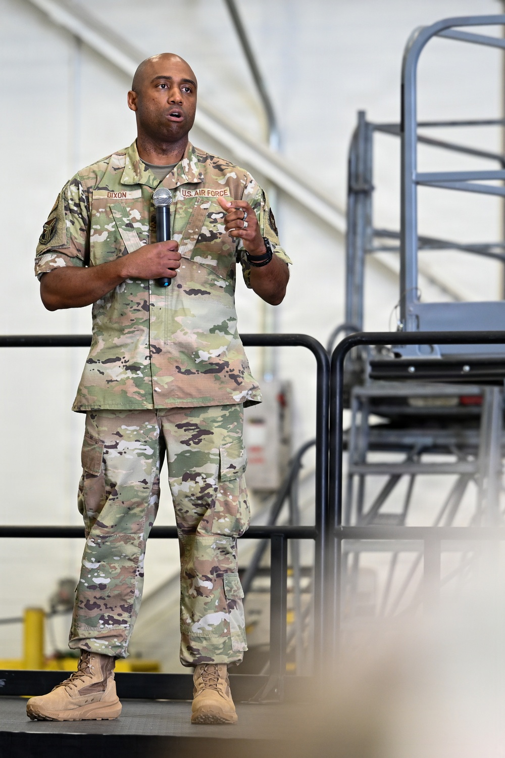 A U.S. Air Force service member named Dixon is standing on a raised platform or stage, holding a microphone and speaking to an audience. He is wearing the Operational Camouflage Pattern (OCP) uniform with 'U.S. AIR FORCE' and 'DIXON' name tapes visible. The setting appears to be inside a military hangar or large facility.