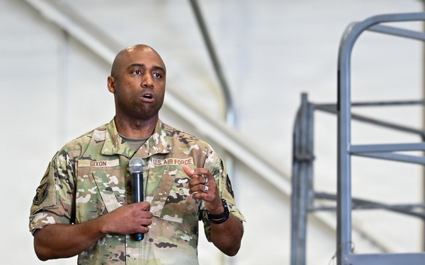 A U.S. Air Force service member named Dixon is standing on a raised platform or stage, holding a microphone and speaking to an audience. He is wearing the Operational Camouflage Pattern (OCP) uniform with 'U.S. AIR FORCE' and 'DIXON' name tapes visible. The setting appears to be inside a military hangar or large facility.
