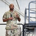 A U.S. Air Force service member named Dixon is standing on a raised platform or stage, holding a microphone and speaking to an audience. He is wearing the Operational Camouflage Pattern (OCP) uniform with 'U.S. AIR FORCE' and 'DIXON' name tapes visible. The setting appears to be inside a military hangar or large facility.