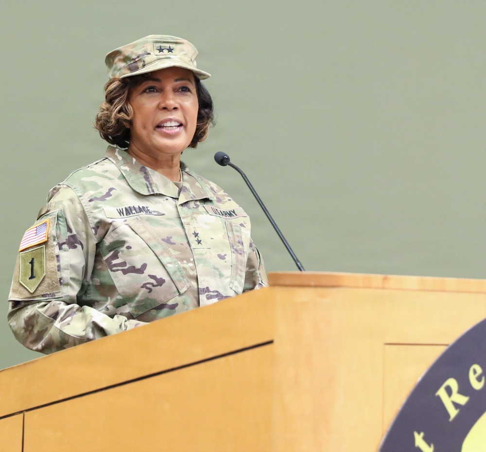 A U.S. Army Major General named Wallace is speaking at a podium during an official military event. She is wearing the Army Combat Uniform (ACU) in Operational Camouflage Pattern (OCP) with two-star rank insignia visible on both her cap and chest. The setting appears to be an indoor military ceremony or briefing.