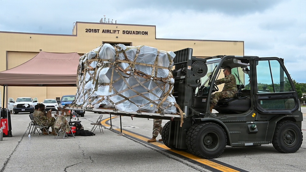 Military personnel are conducting cargo handling operations outside the 201st Airlift Squadron building. A soldier operates a military forklift to move a large, netted and strapped cargo pallet, while other personnel are seated at a table under a canopy tent, likely performing administrative or documentation tasks.