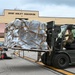 Military personnel are conducting cargo handling operations outside the 201st Airlift Squadron building. A soldier operates a military forklift to move a large, netted and strapped cargo pallet, while other personnel are seated at a table under a canopy tent, likely performing administrative or documentation tasks.