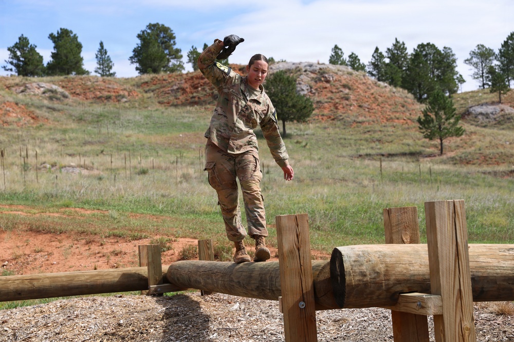 A female soldier in U.S. Army OCP (Operational Camouflage Pattern) uniform is navigating a log balance obstacle on a military obstacle course. She is balancing on a horizontal log supported by wooden posts, with arms extended for stability. The setting appears to be an outdoor training facility in a semi-arid, hilly landscape with pine trees.