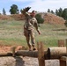 A female soldier in U.S. Army OCP (Operational Camouflage Pattern) uniform is navigating a log balance obstacle on a military obstacle course. She is balancing on a horizontal log supported by wooden posts, with arms extended for stability. The setting appears to be an outdoor training facility in a semi-arid, hilly landscape with pine trees.