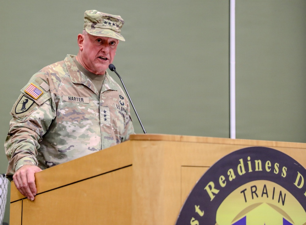 A U.S. Army Lieutenant General named Harter is speaking at a podium during an official military event. He is wearing the Army Combat Uniform (ACU) in MultiCam pattern with three-star rank insignia visible on both his cap and chest. The podium bears a seal referencing 'Readiness' and 'TRAIN,' suggesting a training or readiness-focused military ceremony or briefing.
