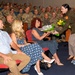 A military ceremony is taking place in an auditorium filled with uniformed soldiers in camouflage. A female officer in an olive/dark green dress uniform is presenting a bouquet of flowers to a seated civilian woman, likely a family member being honored. The audience consists predominantly of military personnel in Army combat uniforms, with a few civilians seated in the front rows.