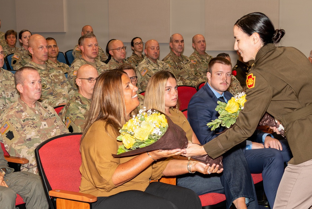 A military ceremony is taking place in an auditorium-style room filled with uniformed U.S. Army personnel seated in red chairs. A female officer in an olive/green uniform is presenting yellow rose bouquets to civilian attendees, likely family members of a service member being honored. The atmosphere appears formal yet warm, consistent with a promotion, retirement, or change-of-command ceremony.