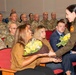 A military ceremony is taking place in an auditorium-style room filled with uniformed U.S. Army personnel seated in red chairs. A female officer in an olive/green uniform is presenting yellow rose bouquets to civilian attendees, likely family members of a service member being honored. The atmosphere appears formal yet warm, consistent with a promotion, retirement, or change-of-command ceremony.