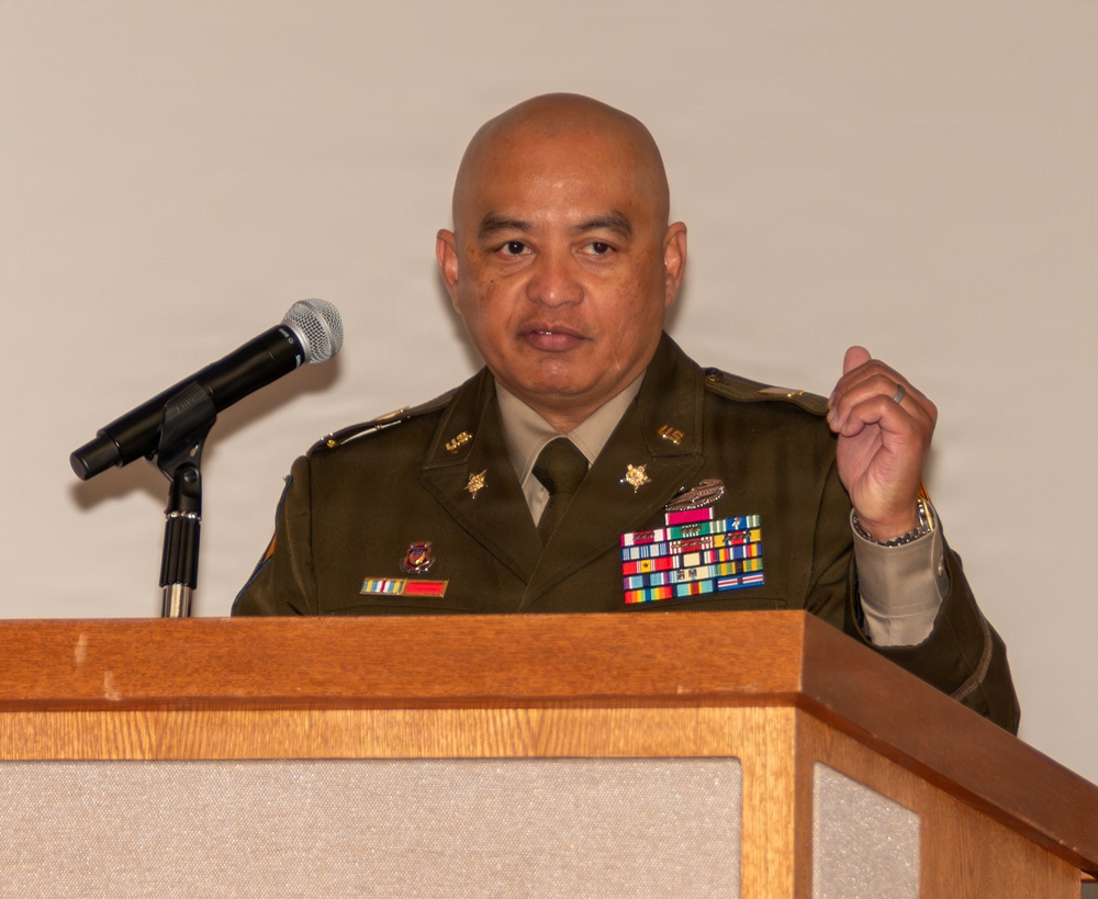 A military officer in dress uniform is speaking at a wooden podium with a microphone. The officer is wearing an olive green Army dress uniform adorned with multiple service ribbons, medals, and insignia. The setting appears to be a formal indoor event or ceremony.