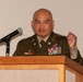 A military officer in dress uniform is speaking at a wooden podium with a microphone. The officer is wearing an olive green Army dress uniform adorned with multiple service ribbons, medals, and insignia. The setting appears to be a formal indoor event or ceremony.