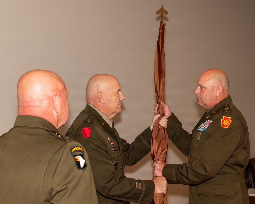 Three military personnel in olive green dress uniforms are participating in a formal guidon or colors transfer ceremony, likely a change of command. Two officers are jointly holding a furled brown/maroon flag on a staff topped with a spade-shaped finial. All three individuals are bald and wear U.S. Army uniforms with visible unit patches.