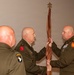 Three military personnel in olive green dress uniforms are participating in a formal guidon or colors transfer ceremony, likely a change of command. Two officers are jointly holding a furled brown/maroon flag on a staff topped with a spade-shaped finial. All three individuals are bald and wear U.S. Army uniforms with visible unit patches.
