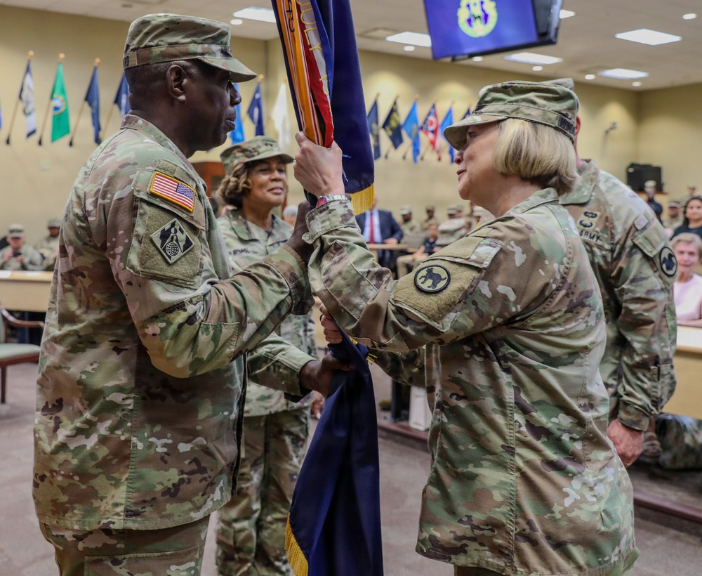 A military change of command ceremony is taking place indoors, with two soldiers in OCP (Operational Camouflage Pattern) uniforms exchanging a unit guidon/colors. The ceremony appears to be conducted in a formal auditorium or conference room setting with an audience of military personnel and civilians in attendance. State and unit flags are displayed along the back wall.