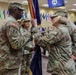 A military change of command ceremony is taking place indoors, with two soldiers in OCP (Operational Camouflage Pattern) uniforms exchanging a unit guidon/colors. The ceremony appears to be conducted in a formal auditorium or conference room setting with an audience of military personnel and civilians in attendance. State and unit flags are displayed along the back wall.