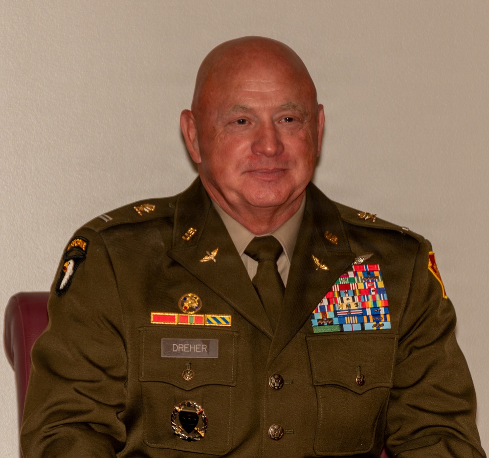 A male U.S. Army soldier in dress green uniform is seated against a plain light-colored wall. His name tag reads 'DREHER' and he displays an extensive array of medals, ribbons, badges, and insignia indicating a long and decorated military career. He appears to be a senior non-commissioned officer or warrant officer based on visible rank and qualification badges.