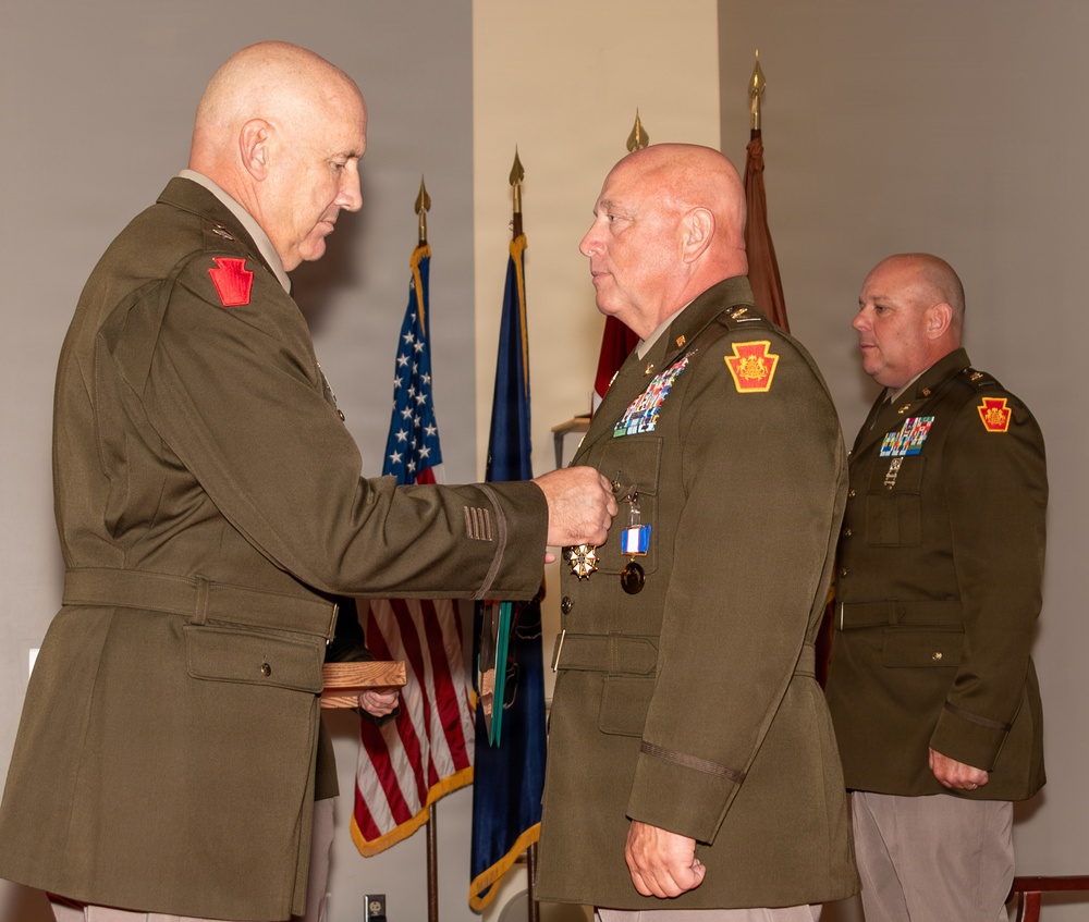 A military decoration ceremony is taking place indoors, where a senior officer is pinning a medal onto the uniform of another officer. Both individuals are wearing olive green Army dress uniforms with Pennsylvania National Guard shoulder patches. A third uniformed officer stands in the background observing the ceremony.