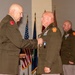 A military decoration ceremony is taking place indoors, where a senior officer is pinning a medal onto the uniform of another officer. Both individuals are wearing olive green Army dress uniforms with Pennsylvania National Guard shoulder patches. A third uniformed officer stands in the background observing the ceremony.