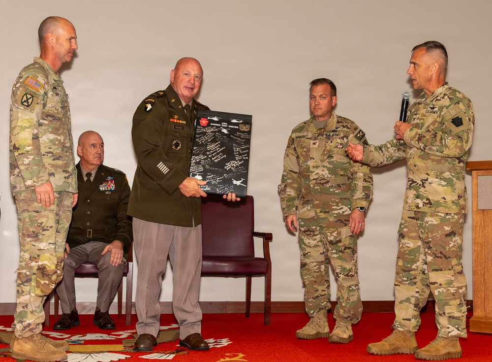 A military ceremony is taking place in what appears to be an indoor venue, likely a retirement or award ceremony. A senior officer in an olive dress uniform is holding a commemorative plaque/shadow box covered in signatures, being presented by fellow soldiers. One soldier is speaking into a microphone while others stand in attendance.