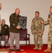 A military ceremony is taking place in what appears to be an indoor venue, likely a retirement or award ceremony. A senior officer in an olive dress uniform is holding a commemorative plaque/shadow box covered in signatures, being presented by fellow soldiers. One soldier is speaking into a microphone while others stand in attendance.