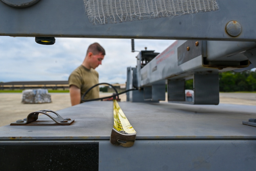 A military service member in an olive drab t-shirt works on or near ground support equipment on what appears to be a military flight line or airfield ramp. The foreground is sharply focused on metal hardware including tie-down hooks and a yellow strap on a gray metal surface, while the person is intentionally blurred in the background. The setting appears to be an outdoor military installation with overcast skies.