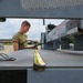 A military service member in an olive drab t-shirt works on or near ground support equipment on what appears to be a military flight line or airfield ramp. The foreground is sharply focused on metal hardware including tie-down hooks and a yellow strap on a gray metal surface, while the person is intentionally blurred in the background. The setting appears to be an outdoor military installation with overcast skies.