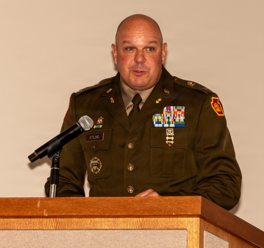 A U.S. Army officer identified by nameplate as 'ETLING' is speaking at a wooden podium with a microphone. He is wearing the Army Green Service Uniform adorned with multiple rows of service ribbons, badges, and insignia. The setting appears to be a formal military ceremony or official function.