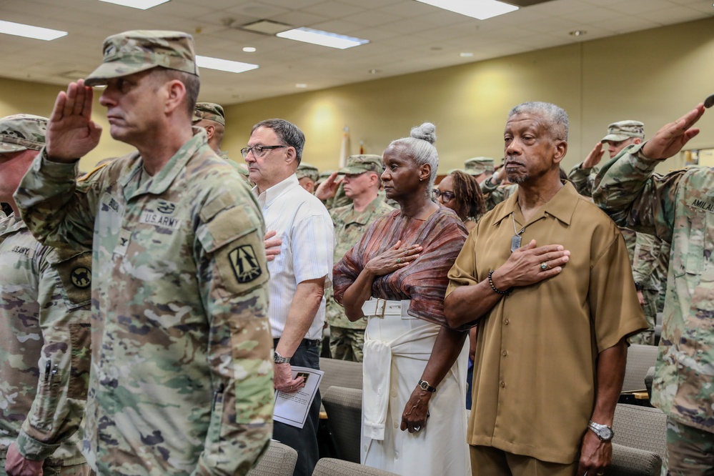 A formal military ceremony is taking place indoors, likely on a U.S. Army installation. Military personnel in Army Combat Uniforms (ACU/OCP pattern) are rendering salutes while civilians stand with their hands over their hearts, suggesting the playing of the National Anthem or Pledge of Allegiance. The mixed group of uniformed soldiers and civilians reflects a formal commemorative or official event.