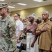 A formal military ceremony is taking place indoors, likely on a U.S. Army installation. Military personnel in Army Combat Uniforms (ACU/OCP pattern) are rendering salutes while civilians stand with their hands over their hearts, suggesting the playing of the National Anthem or Pledge of Allegiance. The mixed group of uniformed soldiers and civilians reflects a formal commemorative or official event.