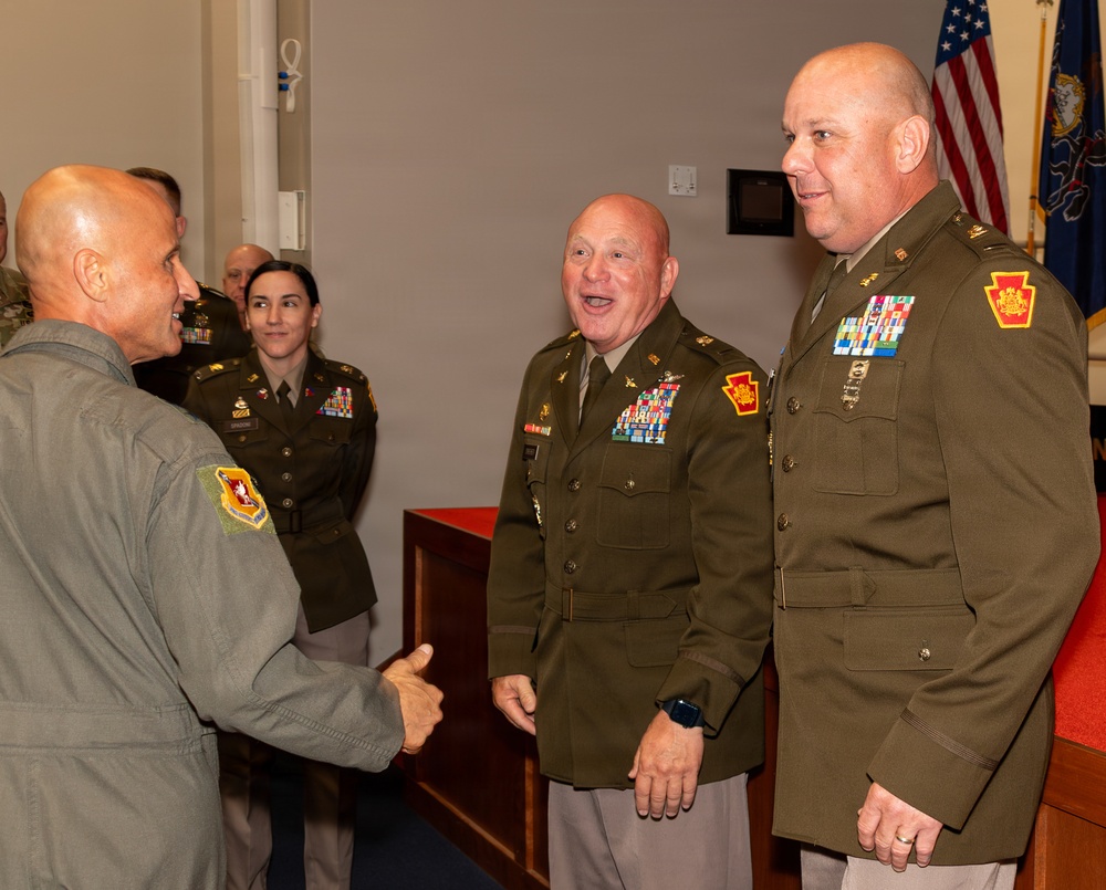 A military ceremony or reception featuring personnel in various uniforms, including Army dress greens and an Air Force flight suit. Two senior Army officers wearing Pennsylvania National Guard shoulder patches and extensive medal/ribbon arrays are engaged in a handshake and conversation with an Air Force officer in a flight suit. Additional uniformed personnel are visible in the background near American and Pennsylvania state flags.