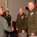 A military ceremony or reception featuring personnel in various uniforms, including Army dress greens and an Air Force flight suit. Two senior Army officers wearing Pennsylvania National Guard shoulder patches and extensive medal/ribbon arrays are engaged in a handshake and conversation with an Air Force officer in a flight suit. Additional uniformed personnel are visible in the background near American and Pennsylvania state flags.
